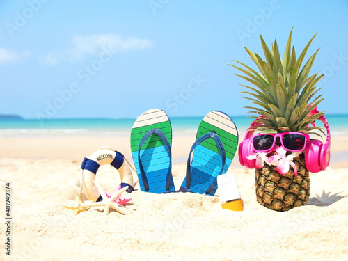 Fototapeta Naklejka Na Ścianę i Meble -  Summer party. Pineapple wearing sunglasses and listen to music with sunblock and sandal on beach and blue sky background. Tropical fashion. Summer Fashion on holiday concept.