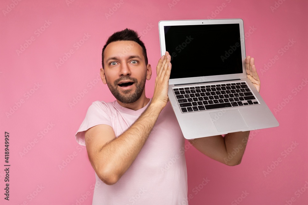 Handsome surprised and amazed man holding laptop computer looking at camera in t-shirt on isolated pink background
