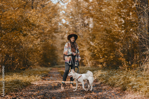 Stylish woman with backpack hiking with white dog in sunny autumn woods. Cute swiss shepherd puppy