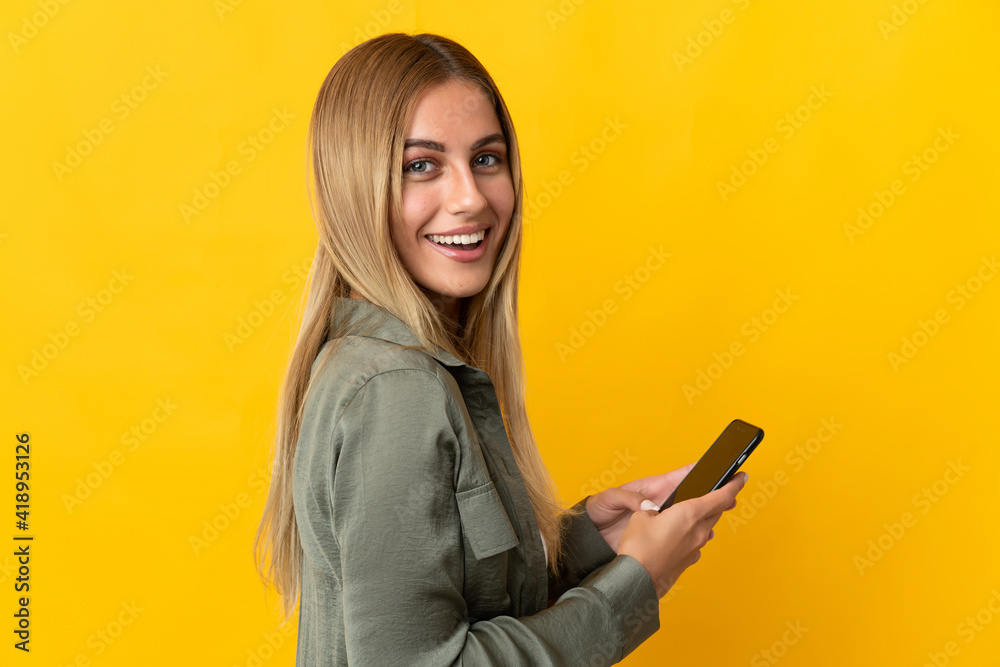 Naklejka premium Young blonde woman isolated on yellow background holding a mobile phone and with arms crossed
