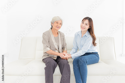 young Asian female holding hand of  old female in home, they feeling happy and smile, they sitting on sofa, mother's day, hand in hand, happiness family time