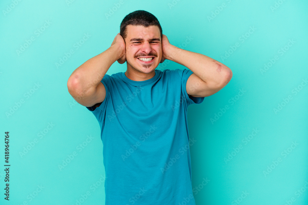 Young caucasian man isolated on blue background covering ears with hands. Young caucasian man isolated on blue background covering ears with hands.