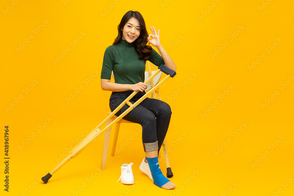 Young Asian woman sitting on chair and sprain foot using crutches and showing ok sign isolated on yellow background, Personal accident concept - obrazy, fototapety, plakaty 