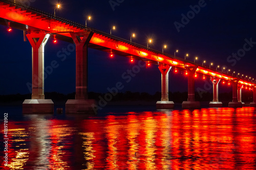 Side view of colourful bridge illuminated with red color lights at the night. Bridge stands on Volga river in Russia. Red light is reflected in the water. © Андрей Рыков