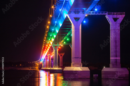 Low angle view of colourful bridge illuminated with different color lights at the night. Bridge stands on Volga river in Russia. Multi-colored light is reflected in the water.