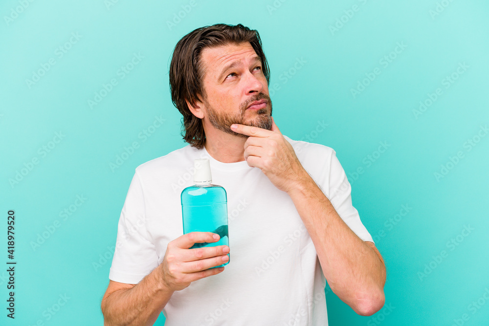 Middle age dutch man sitting holding a mouthwash isolated on blue background looking sideways with doubtful and skeptical expression. Middle age dutch man sitting holding a mouthwash isolated on blue background looking sideways with doubtful and skeptical expression.