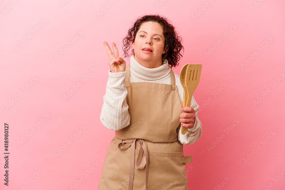 Woman with Down syndrome cooking at home isolated on pink background showing number two with fingers. Woman with Down syndrome cooking at home isolated on pink background showing number two with fingers.