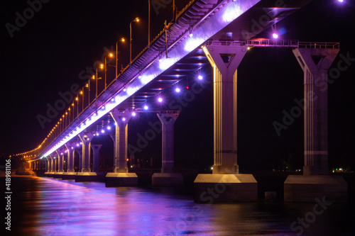 Side view of colourful bridge illuminated with purple color lights at the night. Bridge stands on Volga river in Russia. Purple light is reflected in the water.