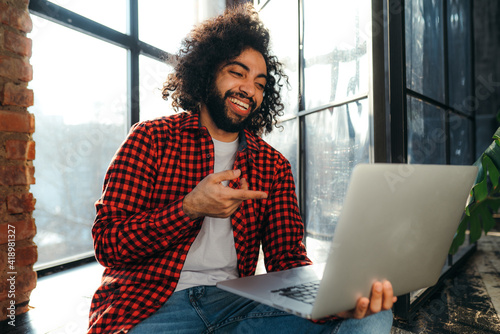Handsome egyptian student in red plaid shirt holding a computer and pointing at it with a finger while laughing
