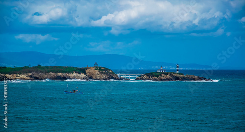 Un faro en una pequeña isla unida a tierra mediante un puente, un barco pesquero de color azul, nubes de tormenta y el mar cantábrico en la costa española