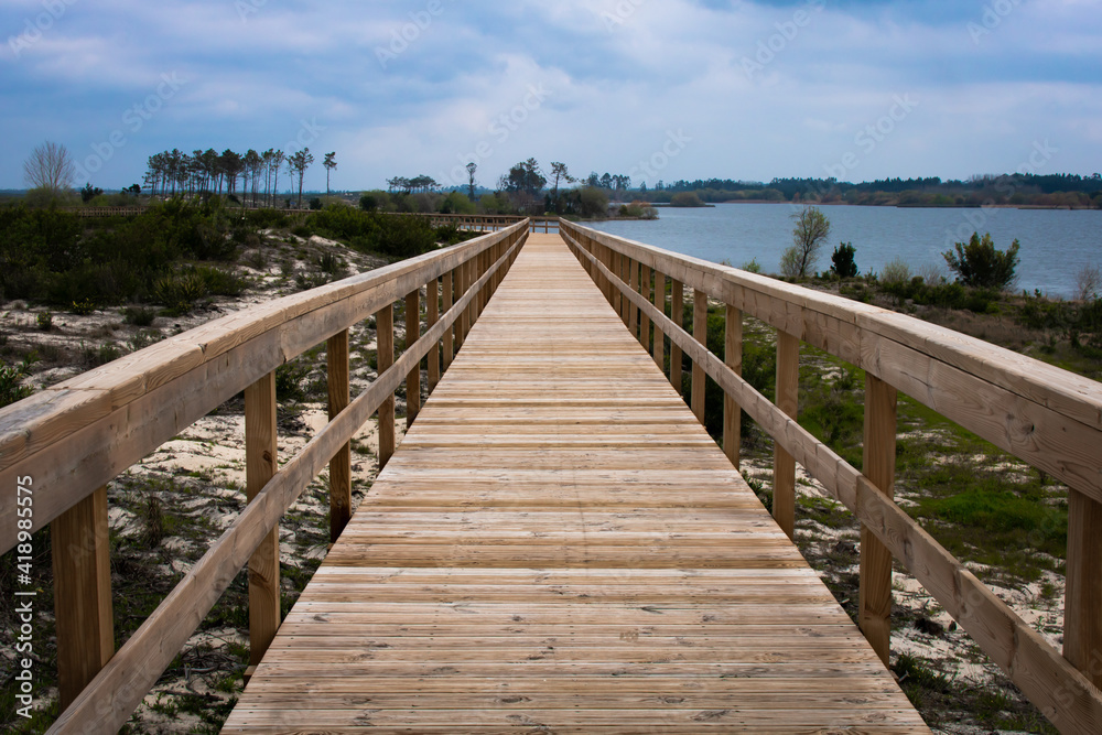 Empty wooden boardwalk and handrail over grassy dune and blue cloudy sky with view over lagoon at Lagoa da Vela, Portugal