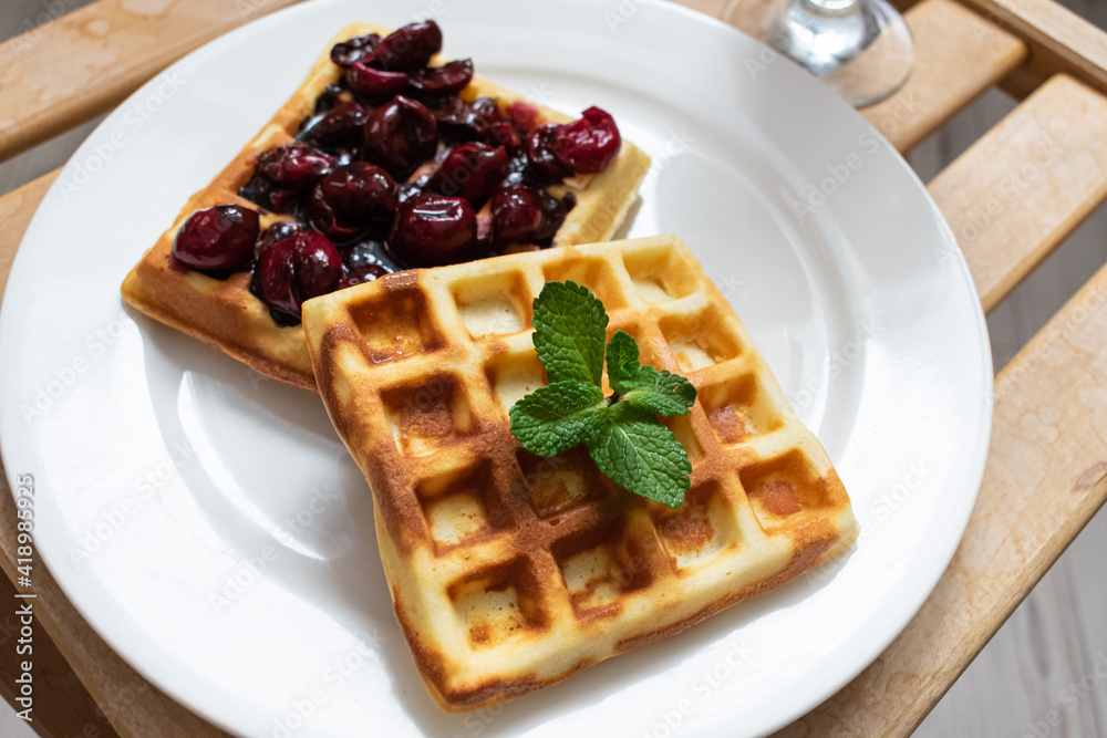 Two Belgian waffles with cherries and mint in a white plate on a wooden tray. Two Belgian waffles with cherries and mint in a white plate on a wooden tray.