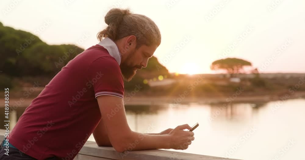 Side view of a man using his gadget while standing on the bridge by the pond. Caucasian guy in red t-shirt texting with sunset skies in the background. Side view of a man using his gadget while standing on the bridge by the pond. Caucasian guy in red t-shirt texting with sunset skies in the background.