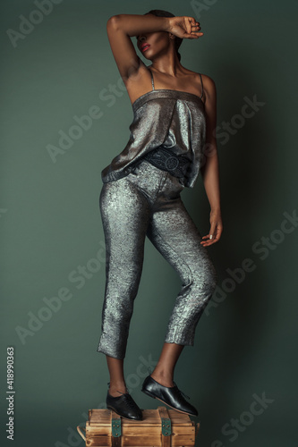 A portrait of a beautiful black woman with long black hair and beautiful makeup posing by herself in a studio on top of a wooden box wearing a silver jumpsuit and black women’s leather lace up shoes.
