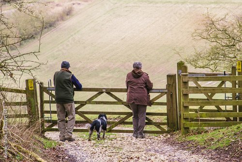 Two walkers and their dog pause to lean on a gate at the top of a hill and admire the view of the countryside below.