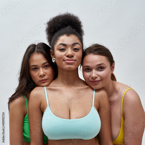 Portrait of three confident female friends, multiethnic young women in colorful underwear looking at camera, posing together isolated over light background