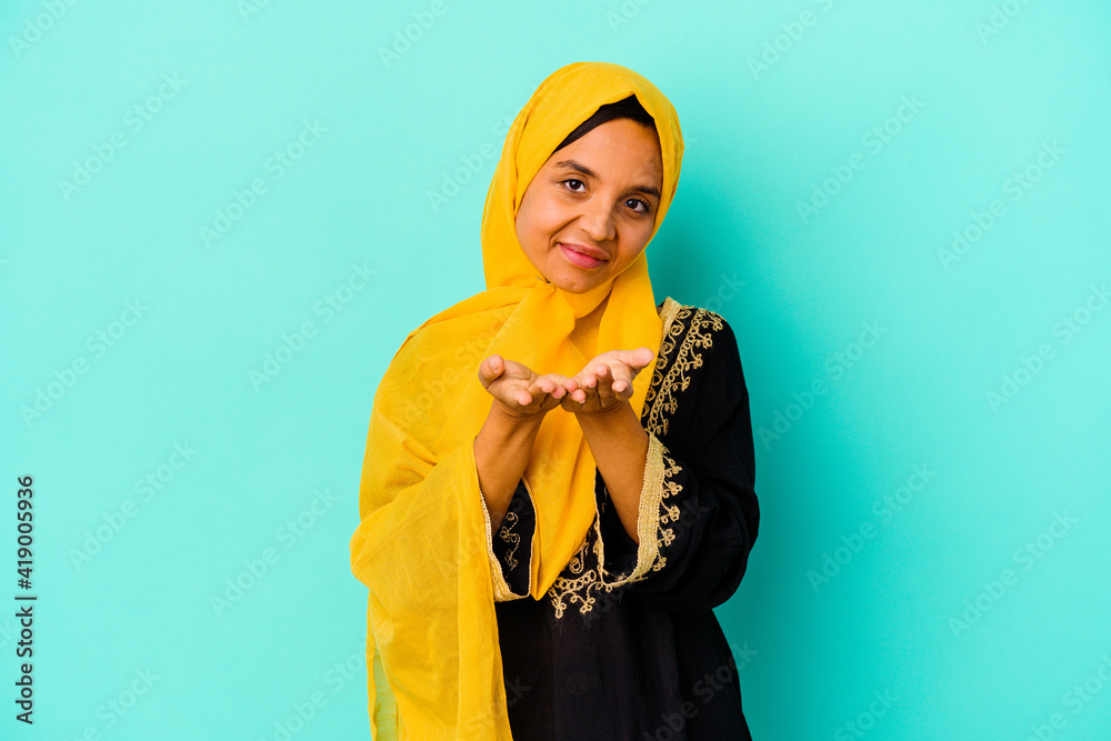 Young muslim woman isolated on blue background holding something with palms, offering to camera. Young muslim woman isolated on blue background holding something with palms, offering to camera.