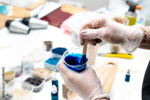 artist's hands in disposable transparent gloves, stirring a blue dye with a wooden spatula in a plastic glass with epoxy resin, in the studio workshop in the background