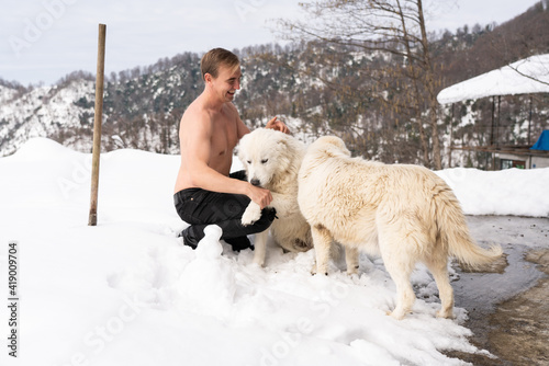 A man with a naked torso caresses a white dog in the snow