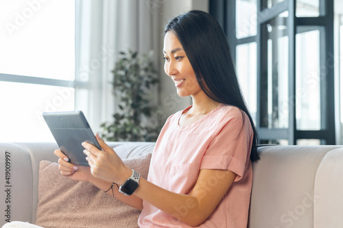 Young Asian mixed-race woman with long black hair sitting on the comfy couch smiling and holding digital tablet, talking with friend online on video call from home, watching comedy show or webinar