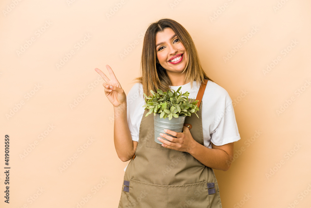 Young indian gardener woman holding a plant isolated joyful and carefree showing a peace symbol with fingers. Young indian gardener woman holding a plant isolated joyful and carefree showing a peace symbol with fingers.