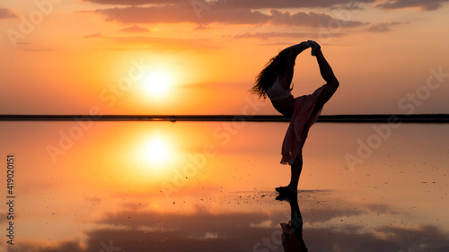 silhouette of a flexible girl in a dress against the background of a golden setting sun reflected in the typing of a graceful woman
