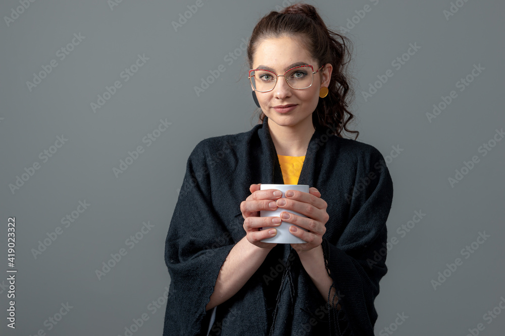 curly-haired emotional young female in a black cloak holds white cup of hot drink with both hands curly-haired emotional young female in a black cloak holds white cup of hot drink with both hands