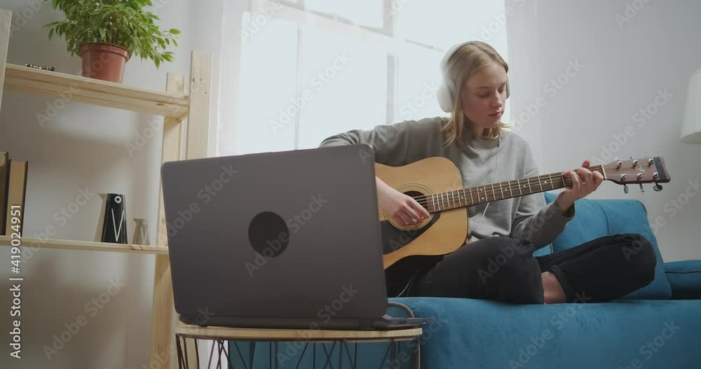 The girl listens to music on headphones using a laptop. A woman is sitting on a blue sofa in a bright living room. The musician selects a melody for the guitar.