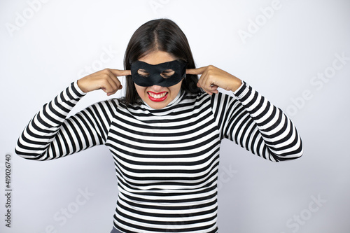 Young beautiful brunette burglar woman wearing mask covering ears with fingers with annoyed expression for the noise of loud music. Deaf concept.