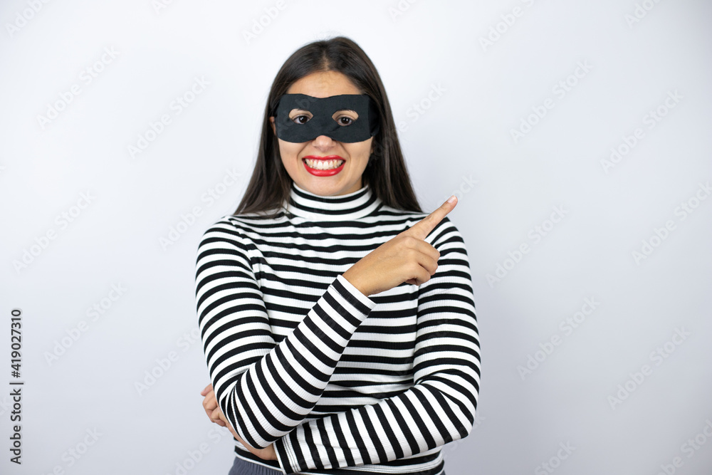 Young beautiful brunette burglar woman wearing mask pointing to the side with a big smile on face