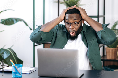 Surprised shocked African American, sits at his desk, holding his head with his hands, looking at a laptop, received an unexpected news or message, saw a financial statement, shocked facial expression