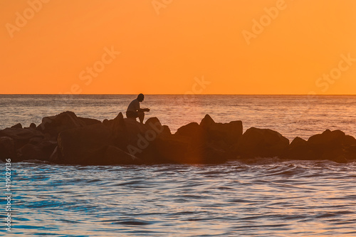 The silhouette of a lonely man sitting in meditation on the rocks of a breakwater near the sea coast against the orange sunset.