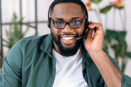 Close-up portrait of an attractive confident bearded African American operator of call center or business leader. Black businessman in headset and stylish shirt looks at the camera and friendly