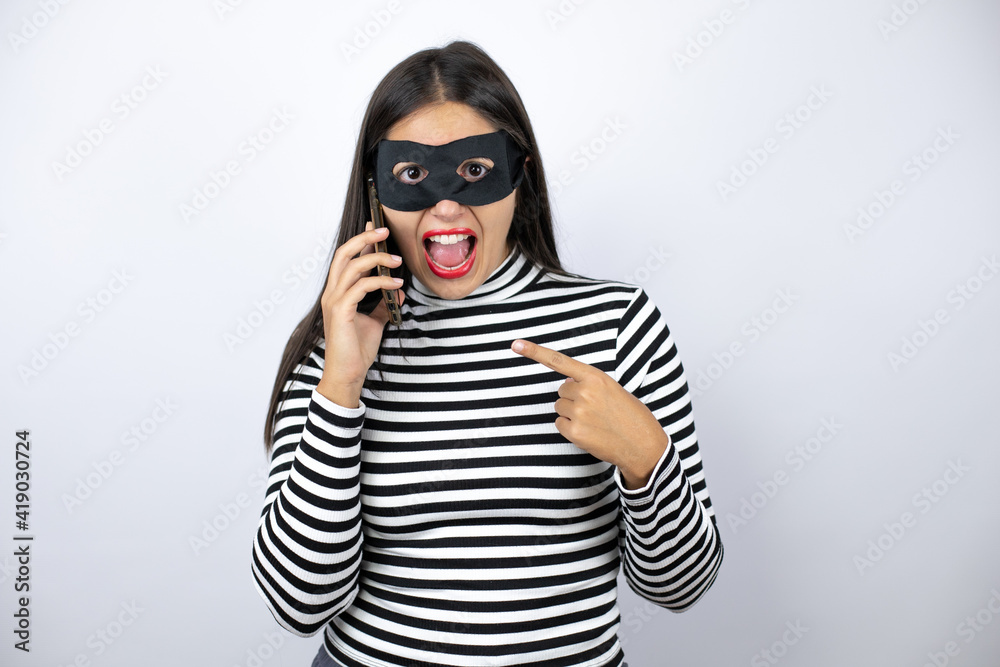 Young beautiful brunette burglar woman wearing mask having conversation talking on the smartphone smiling happy pointing with hand and finger Young beautiful brunette burglar woman wearing mask having conversation talking on the smartphone smiling happy pointing with hand and finger