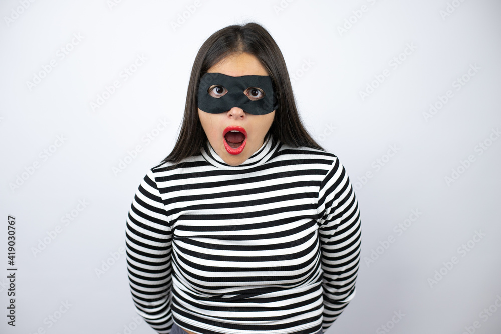Young beautiful brunette burglar woman wearing mask afraid and shocked with surprise and amazed expression, fear and excited face. Young beautiful brunette burglar woman wearing mask afraid and shocked with surprise and amazed expression, fear and excited face.