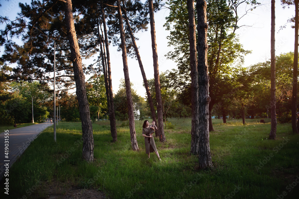 Fototapeta premium Young mother holds baby in arms in the rays of the sunset in the park between pine trees green grass. Woman kisses, hugs girl daughter. Maternal care, custody. Adoption concept. Walks in the open air