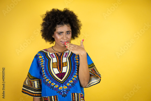 African american woman wearing african clothing over yellow background disgusted with her hand inside her mouth