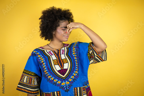 African american woman wearing african clothing over yellow background smelling something stinky and disgusting, intolerable smell, holding breath with fingers on nose