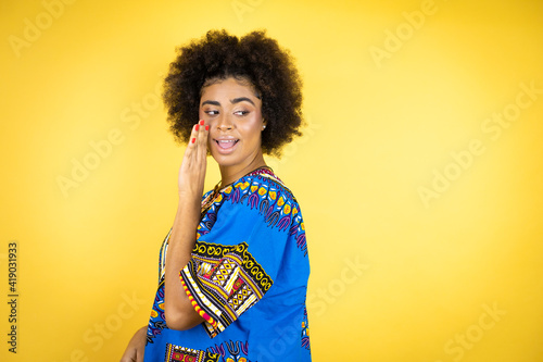 African american woman wearing african clothing over yellow background hand on mouth telling secret rumor, whispering malicious talk conversation