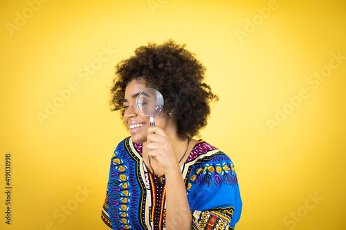 African american woman wearing african clothing over yellow background surprised looking through a magnifying glass