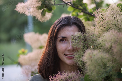 A young woman in a green khaki dress stands by a rosebush Smoky skumpia at sunset in the park. Brunette girl smiling walking in the garden. Close up portrait