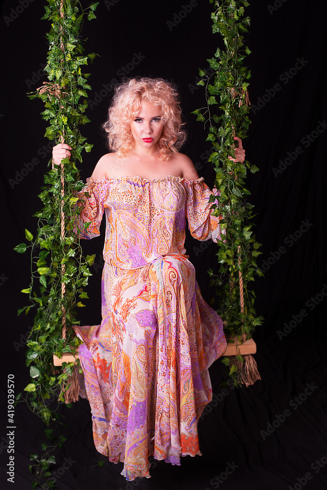 Sexy sad young woman swinging on vintage swing decorated with flowers isolated on black background.