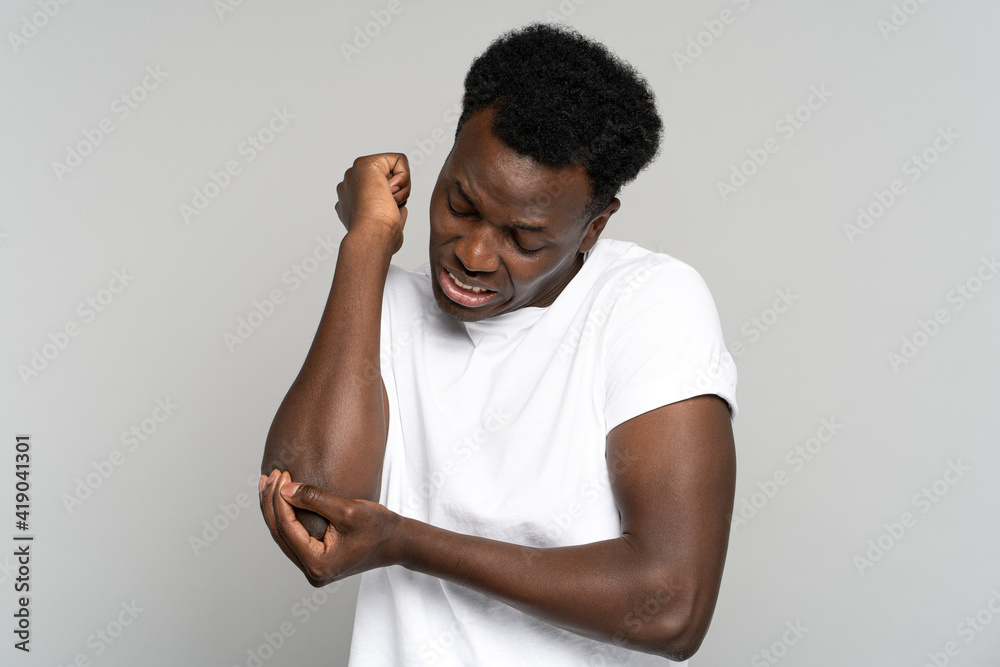 Φωτογραφία Studio portrait of unhappy African American young man in white t-shirt suffers her elbow joint pain or arm bone osteoporosis, pinched nerve, isolated on grey background