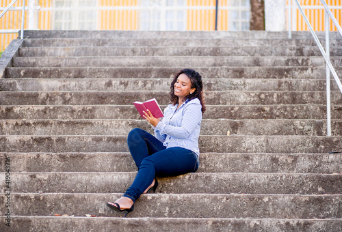 College student sitting on the steps of a college reading a book and smiling.