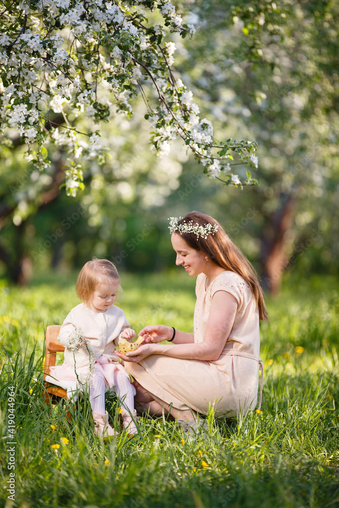 Fototapeta premium Mother and little daughter play in blooming apple garden. Mom loves her child. Spring story. Toddler girl sits on little chair in apple garden. Happy family in beautiful spring day