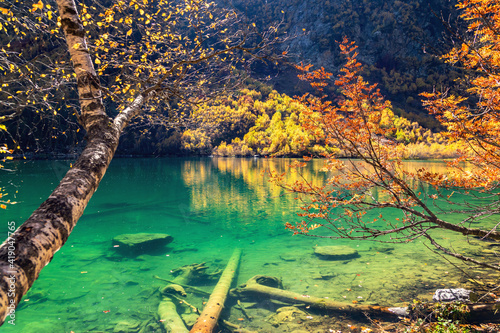 Fototapeta Naklejka Na Ścianę i Meble -  Beautiful landscape with mountain lake and high rocks with illuminated peaks reflection with green water, blue clean sky and yellow autumn sunrise.