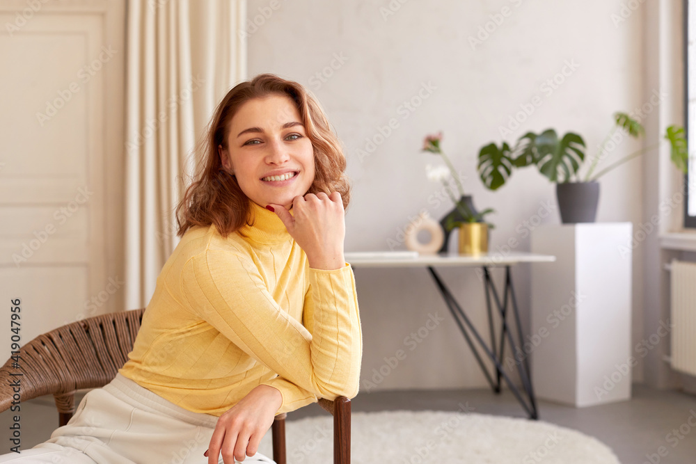 Smiling woman sitting on chair and looking at camera. Content female with charming smile leaning on hand and looking at camera while sitting on wicker chair at home Smiling woman sitting on chair and looking at camera. Content female with charming smile leaning on hand and looking at camera while sitting on wicker chair at home