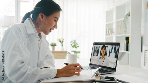 Young Asia lady doctor in white medical uniform using laptop talking video conference call with senior doctor at desk in health clinic or hospital. Social distancing, quarantine for corona virus.