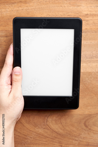 Left hand holding an electronic reader with blank screen on a wooden desk