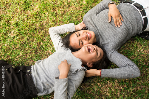 Laughing latin mother elder sister or young baby sitter playing outdoor on the grass with the little girl.